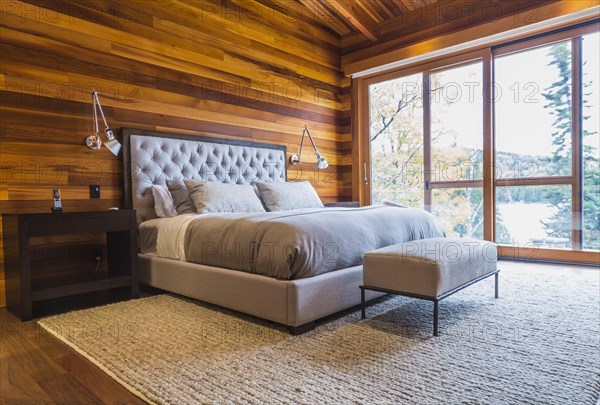 King size bed with grey upholstered panel headboard, ottoman and wooden end tables in master bedroom with grey nuanced rug and Ipe wood floor inside luxurious stained cedar and timber wood home with panoramic windows, Quebec, Canada, North America