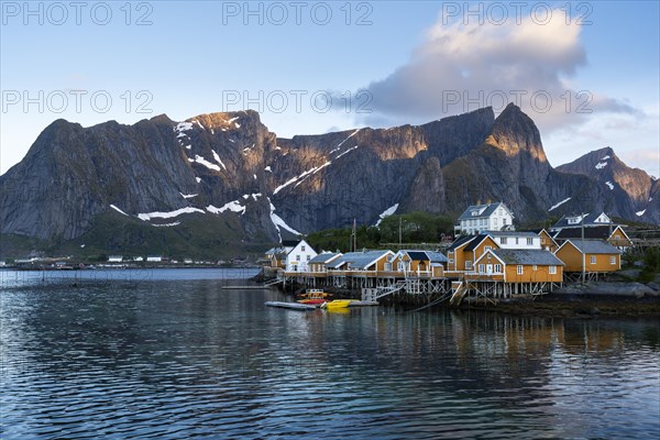 The village of Sakrisoy with its typical yellow or ochre-coloured wooden houses on wooden stilts (rorbuer) by the sea. At night at the time of the midnight sun in good weather. Blue sky, one cloud. Sunlight on the mountain peaks. Early summer. Sakrisoy, Moskenesoya, Lofoten, Norway, Europe