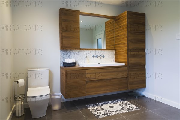 White high-back flush toilet and American walnut wood vanity with white rectangular sink and ceramic tile flooring in main bathroom on upstairs floor inside modern cube style home, Quebec, Canada, North America