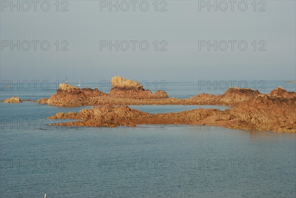 A serene seascape with a rocky shoreline and sailing boats in the distance during golden hour