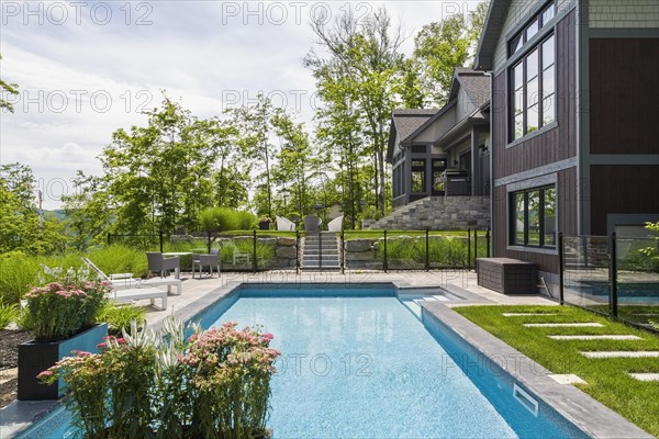 In-ground swimming pool and rear view of contemporary natural stone and brown stained wood and cedar shingles clad luxurious bungalow style home in summer, Quebec, Canada, North America