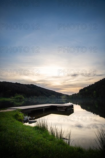 A lake in a landscape shot. A sunset and the natural surroundings are reflected in the water of the reservoir. Marbach reservoir, Odenwald, Hesse