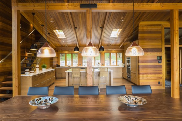 Illuminated industrial style copper with frosted glass pendant lighting fixtures over wooden dining table with black leather chairs plus kitchen with bamboo wood island and cabinets in background inside luxurious stained cedar and timber wood home with panoramic windows, Quebec, Canada, North America