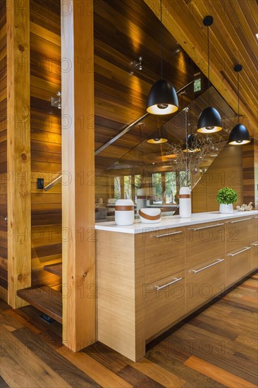 Bamboo wood buffet with white quartz countertop illuminated by black industrial style pendant lighting fixtures next to staircase with clear glass railing in kitchen with Ipe wood floor inside luxurious stained cedar and timber wood home, Quebec, Canada, North America