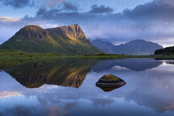 Landscape on the Lofoten Islands. The mountain Offersoykammen and the bay Vagspollen. Mount Stornappstinden on Flakstadoya in the background. The mountains are reflected in the water. At night during the midnight sun. A few clouds in the sky. The night sun shines on the mountain peak. Golden hour. Early summer. Vestvagoya, Lofoten, Norway, Europe