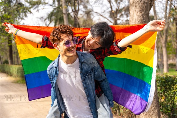 Portrait of a joyful multi-ethnic gay couple waving lgbt flag piggybacking in a park