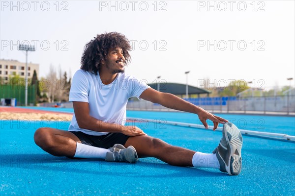 Side view of a smiling young african sportive man stretching sitting on a running track