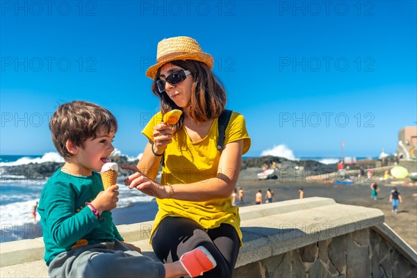 A woman and a child are sitting on a wall near the ocean, eating ice cream. The woman is wearing a yellow shirt and a straw hat. Scene is lighthearted and fun, as the two enjoy their time together