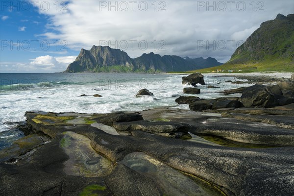 Seascape on the beach at Uttakleiv (Utakleiv), in the foreground rocks and rocky outcrops filled with water, including the so-called Eye of Uttakleiv, a round stone in the water. Mount Hogskolmen in the background. Sun and clouds. Early summer. Uttakleiv, Vestvagoya, Lofoten, Norway, Europe