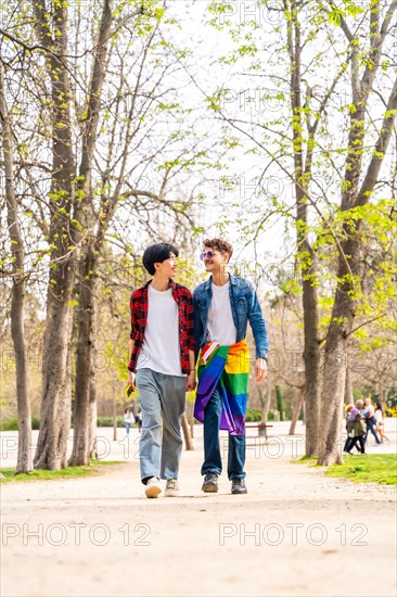 Vertical full length photo of a gay male couple walking along a park carrying LGBT rainbow flag