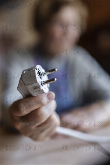 Senior citizen holding a power cable with plug in her hand at home, symbolising energy costs and poverty, Cologne, North Rhine-Westphalia, Germany, Europe