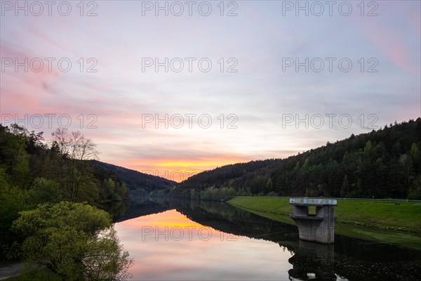 A lake in a landscape shot. A sunset and the natural surroundings are reflected in the water of the reservoir. Marbach reservoir, Odenwald, Hesse