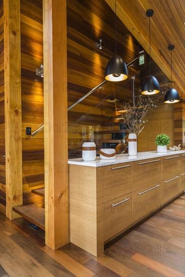 Bamboo wood buffet with white quartz countertop illuminated by black industrial style pendant lighting fixtures next to staircase with clear glass railing in kitchen with Ipe wood floor inside luxurious stained cedar and timber wood home, Quebec, Canada, North America