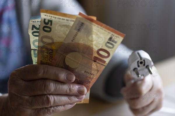 Senior citizen holding money and a power cable with plug in her hand at home, symbolising energy costs and poverty, Cologne, North Rhine-Westphalia, Germany, Europe