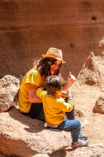 A woman and a child are sitting on a rock in a desert. The woman is wearing a yellow shirt and a straw hat. They are both smiling and seem to be enjoying their time together