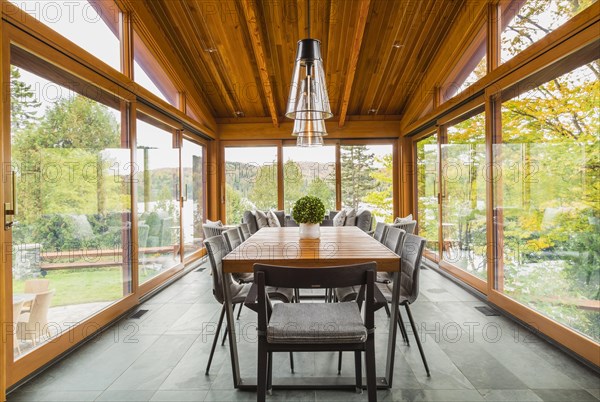 Four season gazebo and illuminated industrial style black with clear glass pendant lighting fixtures over wood and black metal dining table with grey upholstered chairs and heated slate floor inside luxurious stained cedar and timber wood home with panoramic windows, Quebec, Canada, North America