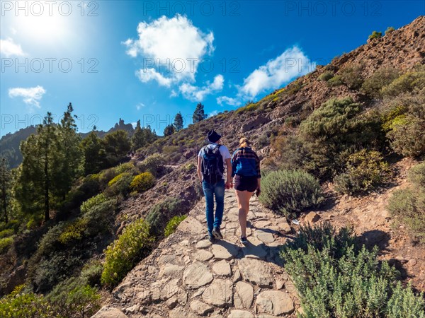 A man and woman are walking on a path in the woods. The woman is wearing a backpack and the man is wearing a hat. The sky is blue and there are trees in the background
