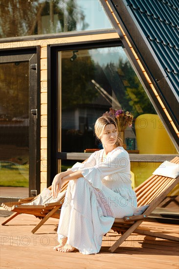 Woman sitting on wooden folding chair on terrace of a-frame cabin and looking to the side