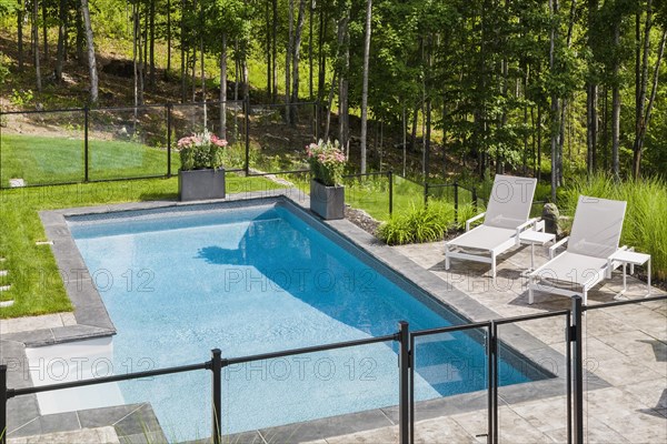 Two white long chairs on edge of in-ground swimming pool enclosed by clear glass and black metal fence in residential backyard in summer, Quebec, Canada, North America