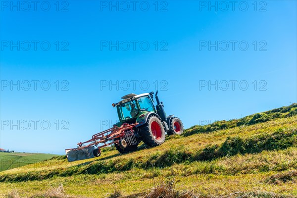 A tractor is driving down a hillside, plowing the grass. The sky is clear and blue, and the sun is shining brightly. The scene is peaceful and serene, with the tractor moving slowly