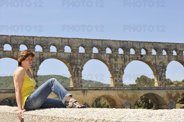 Woman sitting on a wall in front of the Pont du Gard, Roman aqueduct over the river Gardon, Vers-Pont-du-Gard, Languedoc-Roussillon, South of France, France, Europe