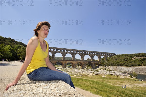 Woman sitting on a wall in front of the Pont du Gard, Roman aqueduct over the river Gardon, Vers-Pont-du-Gard, Languedoc-Roussillon, South of France, France, Europe