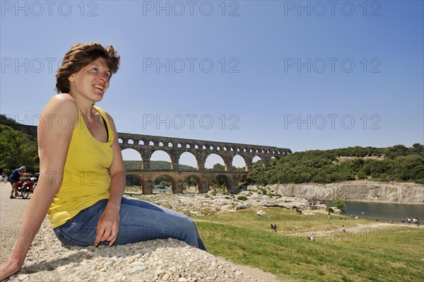Woman sitting on a wall in front of the Pont du Gard, Roman aqueduct over the river Gardon, Vers-Pont-du-Gard, Languedoc-Roussillon, South of France, France, Europe