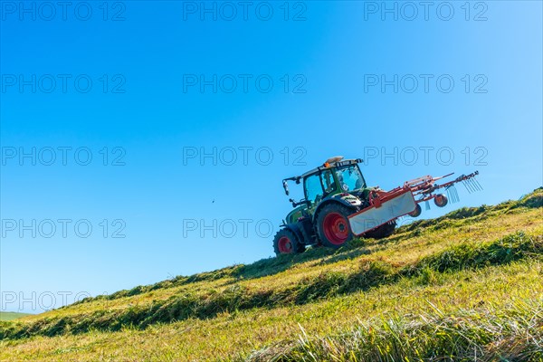 A tractor is driving down a hill, plowing the grass. The sky is clear and blue, and there are no clouds in sight