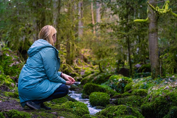 A woman sits on the bank of a forest stream, surrounded by moss and green vegetation, and seems to be thinking, Calw, Black Forest, Germany, Europe