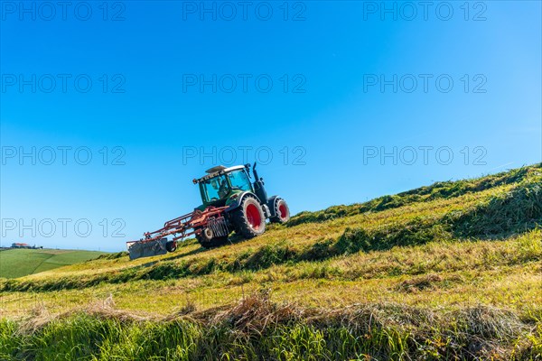 A tractor is driving down a hillside, plowing the grass. The sky is clear and blue, and the sun is shining brightly. The scene is peaceful and serene, with the tractor moving slowly