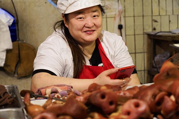 Chongqing, Chongqing Province, China, A smiling vendor with a red apron holds a mobile phone in a market stall full of pig heads, Chongqing, Chongqing, Chongqing Province, China, Asia