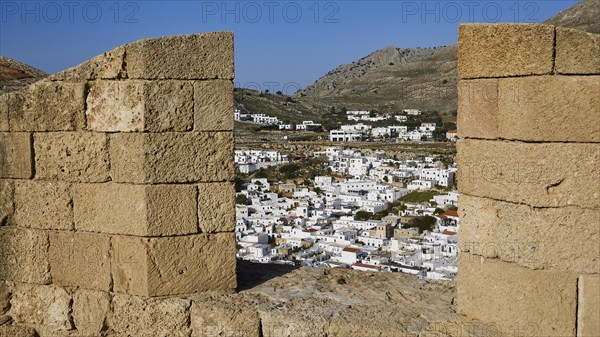 View through an old stone wall to a settlement with white houses and mountains in the background, St John's Fortress, Lindos, Rhodes, Dodecanese, Greek Islands, Greece, Europe