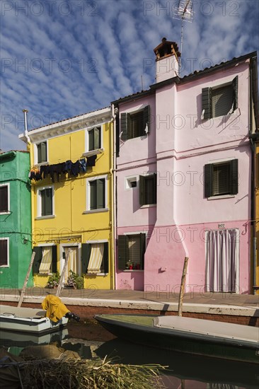 Moored boats on canal lined with pink, yellow and green stucco houses decorated with striped curtains over entrance doors and windows plus clothes on clothesline, Burano Island, Venetian Lagoon, Venice, Veneto, Italy, Europe