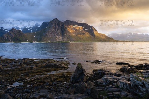 Landscape with sea and mountains on the Lofoten Islands, view across the fjord to the small town of Flakstad and the mountain Flakstadtinden as well as other mountains. Rocks in the foreground. At night at the time of the midnight sun. Cloudy sky. The night sun shines on the mountain peak. Early summer. Golden hour. Flakstadoya, Lofoten, Norway, Europe