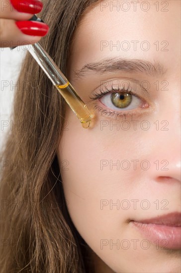 Happy young woman applying serum on her face with her eyes open. Beautiful young Brazilian woman moisturizes her skin with serum isolated against white background