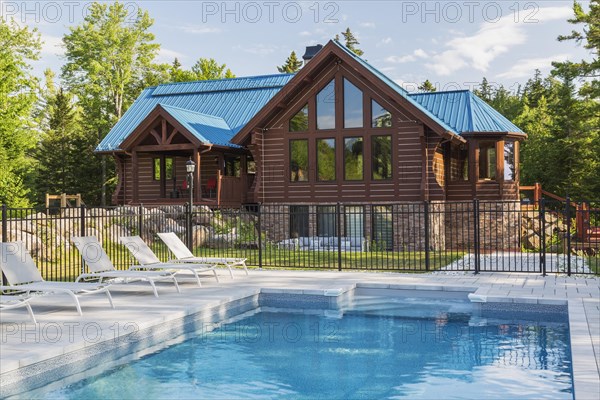 In-ground salt water swimming pool and brown stained milled Eastern white pine timber and flat log profile home facade with stone cladding on walk-out lower level and blue standing-seam sheet metal roof, Quebec, Canada, North America