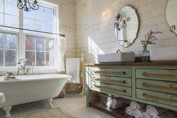 Green antique finish pine wood vanity with rustic wood plank countertop and white top-mounted his and hers vessel sinks, freestanding claw foot bathtub in en suite with honeycomb ceramic tile flooring on upstairs floor inside country style home, Quebec, Canada, North America