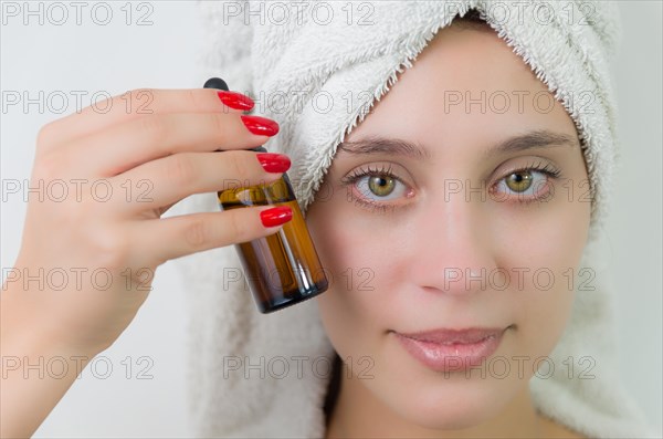 Happy young woman with towel on head holding amber glass bottle of skin serum. Beautiful young Brazilian woman moisturizes her skin with serum isolated against white background