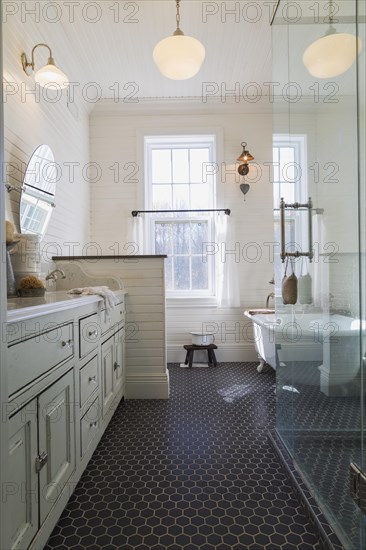White antique style finish vanity with quartz countertop and two sunken sinks, freestanding claw foot bathtub, glass shower stall in kid's upstairs floor bathroom with black honeycomb ceramic tile floor inside country style home, Quebec, Canada, North America