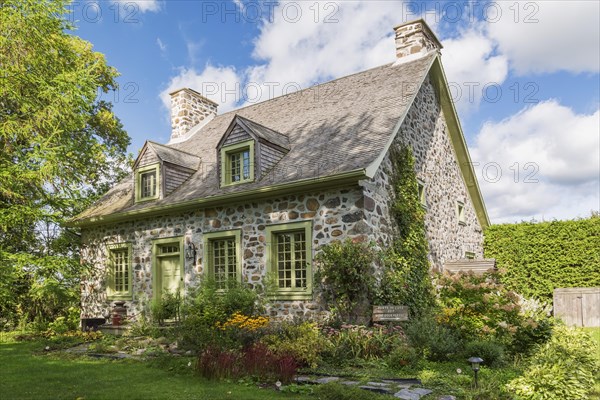 Old circa 1735 Canadiana fieldstone house facade with lime green trim and cedar wood shingles roof and landscaped front yard in late summer, Quebec, Canada, North America