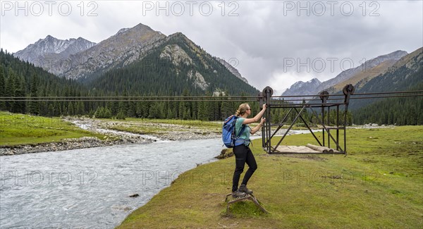 Hiker with backpack standing in front of a drawbridge in a mountainous landscape by the river, Chong Kyzyl Suu Valley, Terskey Ala Too, Tien-Shan Mountains, Kyrgyzstan, Asia