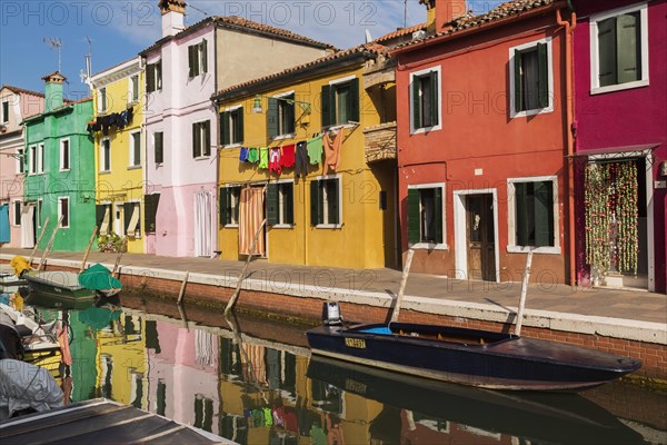 Moored boats on canal lined with pink, red, orange, yellow and green stucco houses decorated with curtains over entrance doors plus washed clothes on clothesline, Burano Island, Venetian Lagoon, Veneto, Italy, Europe