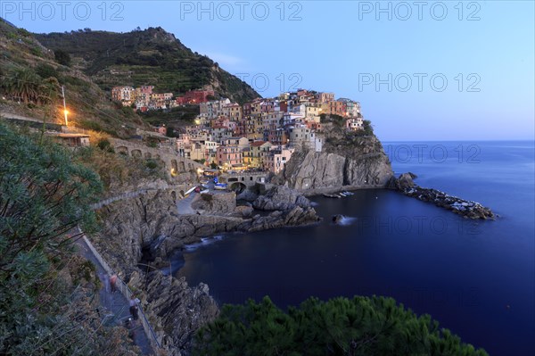 Mediterranean village on rocks on the coast at dusk with lights switched on and calm sea, Italy, Liguria, Manarola, Riomaggiore, La Spezia Province, Cinque Terre, Europe