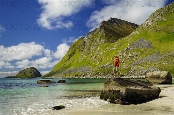 Landscape with sea at the sandy beach of Haukland (Hauklandstranda) with the mountain Veggen. A woman stands barefoot on a rock and looks out to sea. Shot during the day with blue sky. Early summer. Haukland Beach, Haukland, Vestvagoya, Lofoten, Norway, Europe