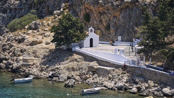 A white church on the Mediterranean Sea on a rocky shore next to parked boats, St Paul's Chapel, Paul's Bay, below the Acropolis of Lindos, Lindos, Rhodes, Dodecanese, Greek Islands, Greece, Europe