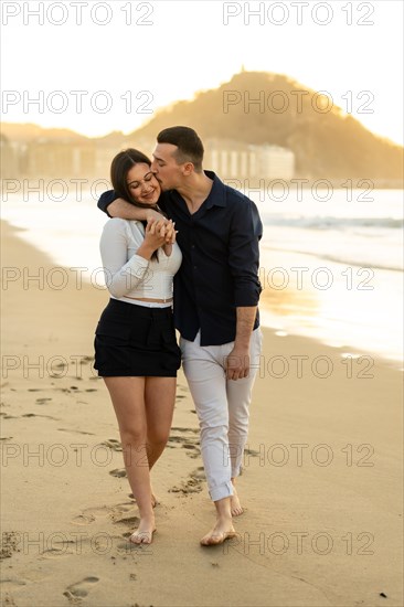 Vertical photo of a caucasian young couple kissing while walking barefoot along a beach during sunset