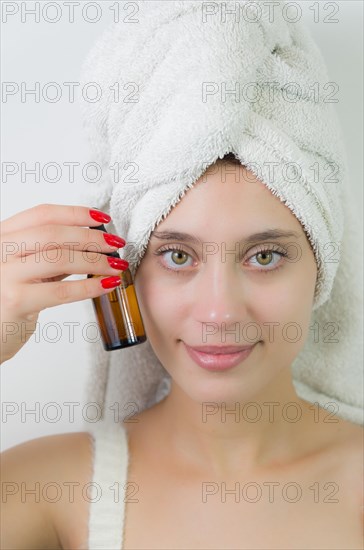 Happy young woman with towel on head holding amber glass bottle of skin serum. Beautiful young Brazilian woman moisturizes her skin with serum isolated against white background