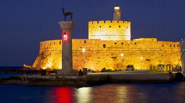 Illuminated historic fortress by the sea at dusk with clear sky and lighthouse, night shot, European roe deer statue, Fort of Saint Nikolaos, harbour promenade, Rhodes, Dodecanese, Greek Islands, Greece, Europe