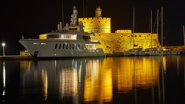 Night scene of an illuminated castle and a lighthouse with reflections near a yacht, night shot, Fort of Saint Nikolaos, harbour promenade, Rhodes, Dodecanese, Greek Islands, Greece, Europe