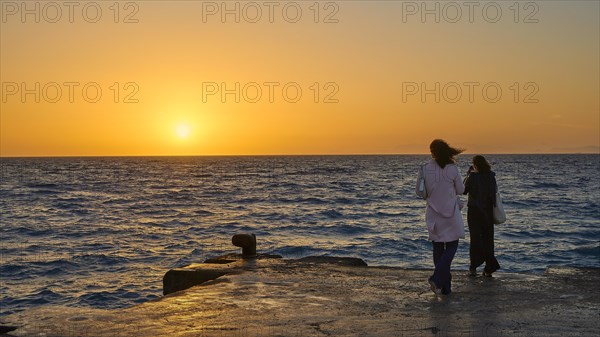 A couple stands on the shore chatting with a view of the sun setting over the sea, dusk, sunset, Western Promenade, Rhodes Town, Rhodes, Dodecanese, Greek Islands, Greece, Europe